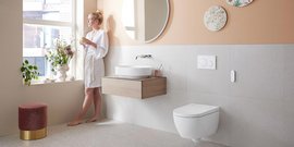 Eine Frau in einem weißen Bademantel steht am Fenster und genießt ein Getränk, umgeben von Pflanzen und einem modernen Badezimmer.