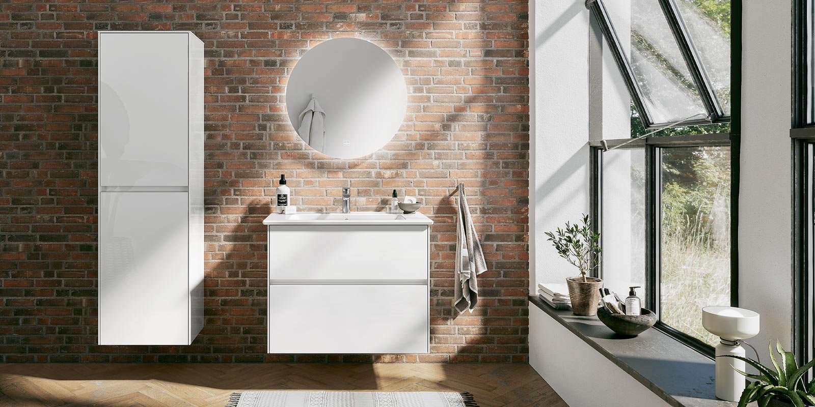 Ein modernes Badezimmer mit einer weißen Waschtischkommode und einem runden Spiegel vor einer Backsteinwand.